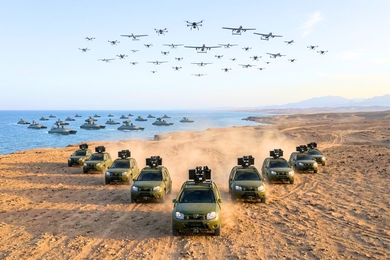 A swarm of small drones flying over a formation of unmanned ground vehicles on a beach beside the sea